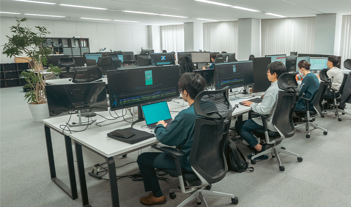 デュアルモニターと高機能チェアが並ぶ執務フロアで作業するメンバー